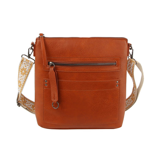The Daily Zip Pocket Guitar Strap Crossbody Bag is a brown faux leather bag with silver-tone hardware, a front zip pocket, decorative key ring, and an adjustable beige strap with a white geometric pattern.