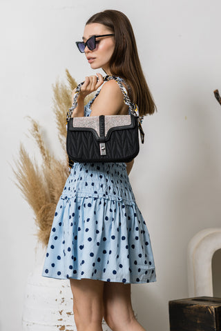 A woman in sunglasses and a light blue polka dot dress poses indoors, holding the Scarf Accented Sparkling Shoulder Bag, featuring a textured strap and silver detail, next to dried plants and a white chair.