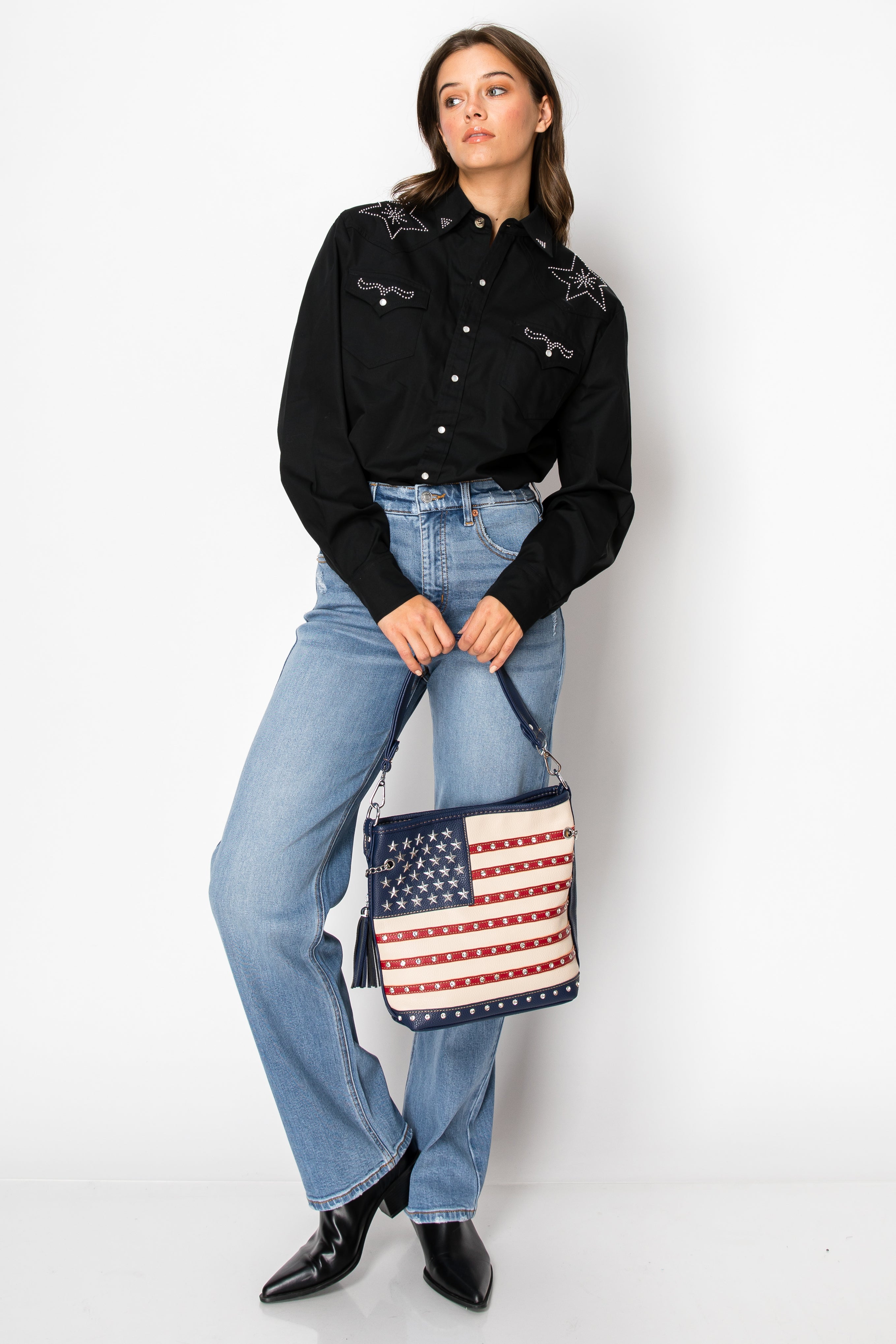 A woman in a black star-detailed button-up, light blue jeans, and black boots poses with the American Flag Hobo Crossbody against a plain white background.