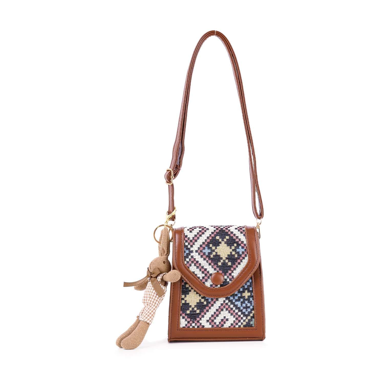 The Decorative Accent Shoulder Bag is a small, lightweight brown crossbody with an adjustable strap, a colorful geometric tapestry flap in blue, white, red, and yellow, and a plush bunny keychain on the side.