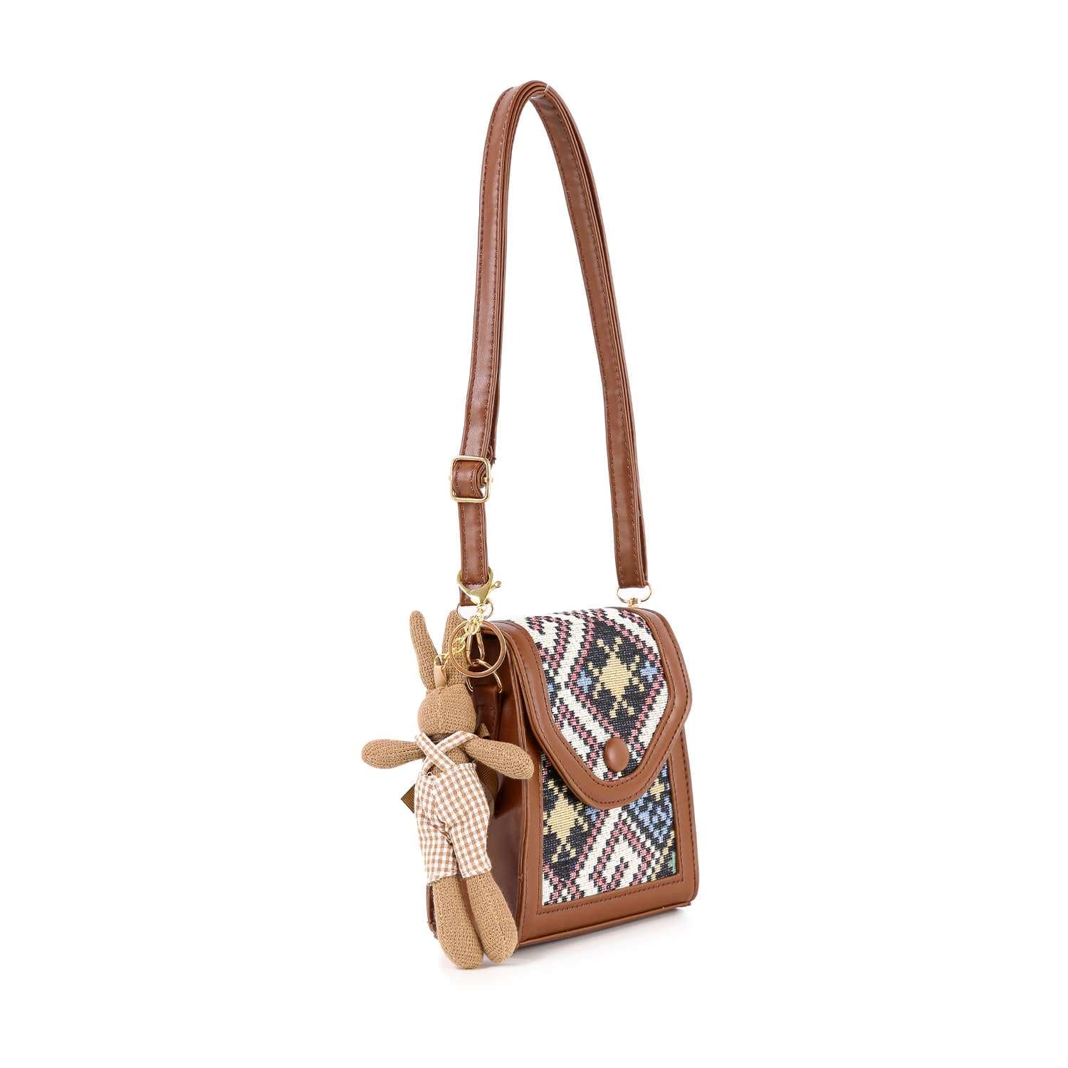The Decorative Accent Shoulder Bag is a brown lightweight crossbody with a tapestry flap and front in dark blue, white, and beige geometric patterns. It includes an adjustable strap and a checkered outfit bear keychain.