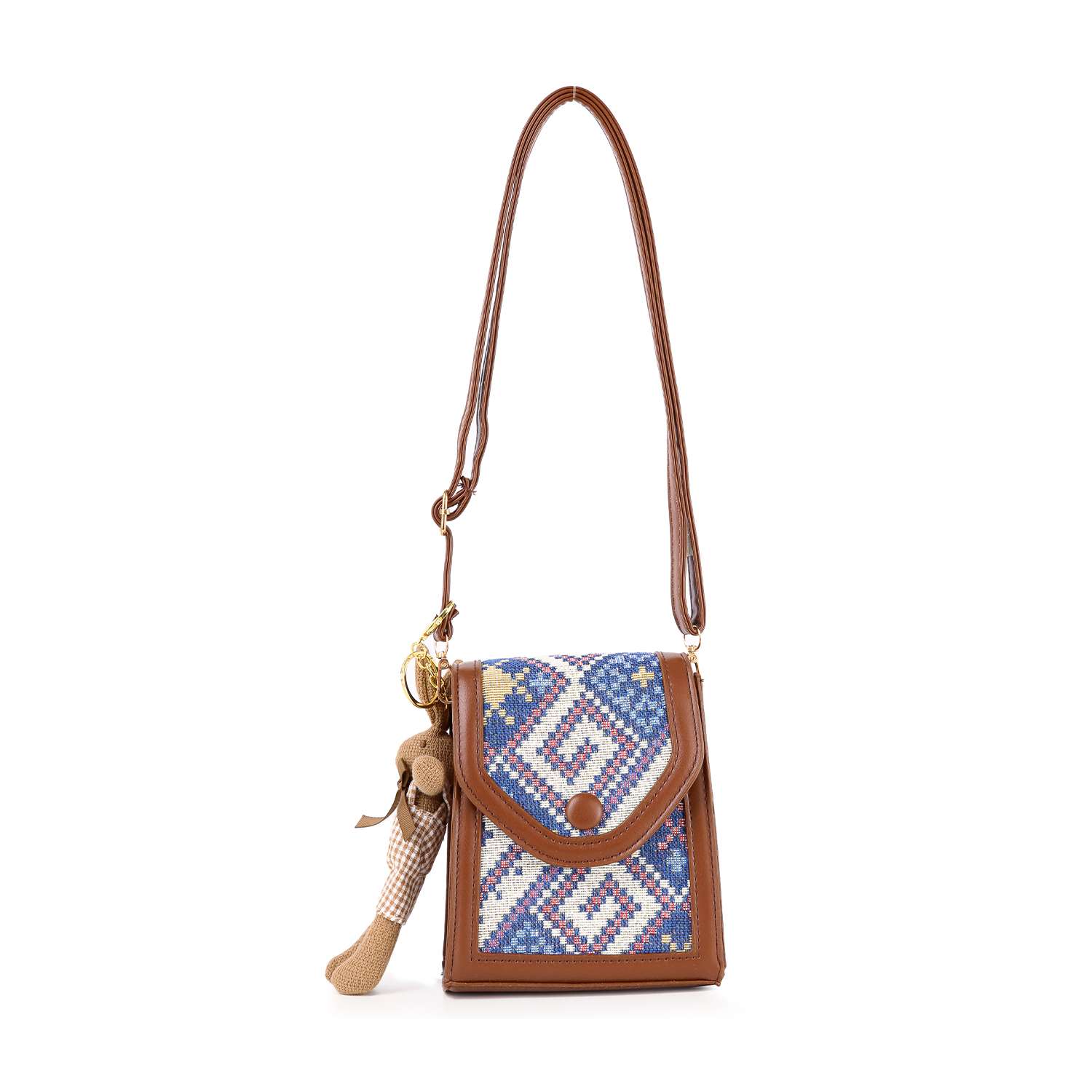 The Decorative Accent Shoulder Bag is a lightweight crossbody with an adjustable strap, showcasing a blue, white, and purple tapestry flap. It includes a beige and brown plush keychain attached to the side.