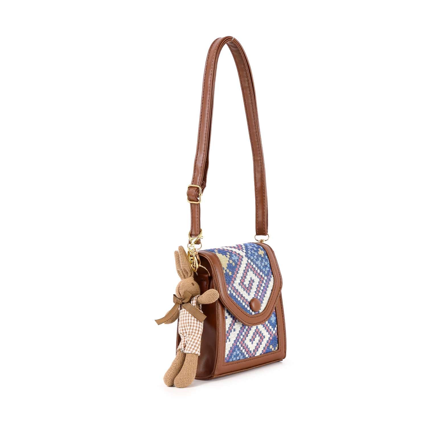 The Decorative Accent Shoulder Bag is a lightweight brown crossbody with an adjustable strap, featuring a blue and white geometric tapestry flap and a plush bunny keychain on the side.