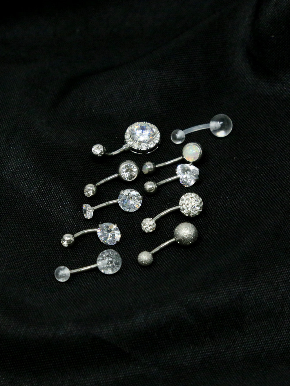 The Set of 10 Dazzling Belly Button Rings in 8mm sizes is displayed on black fabric, featuring crystal, glitter, and opal-like designs—perfect styles for everyday wear.