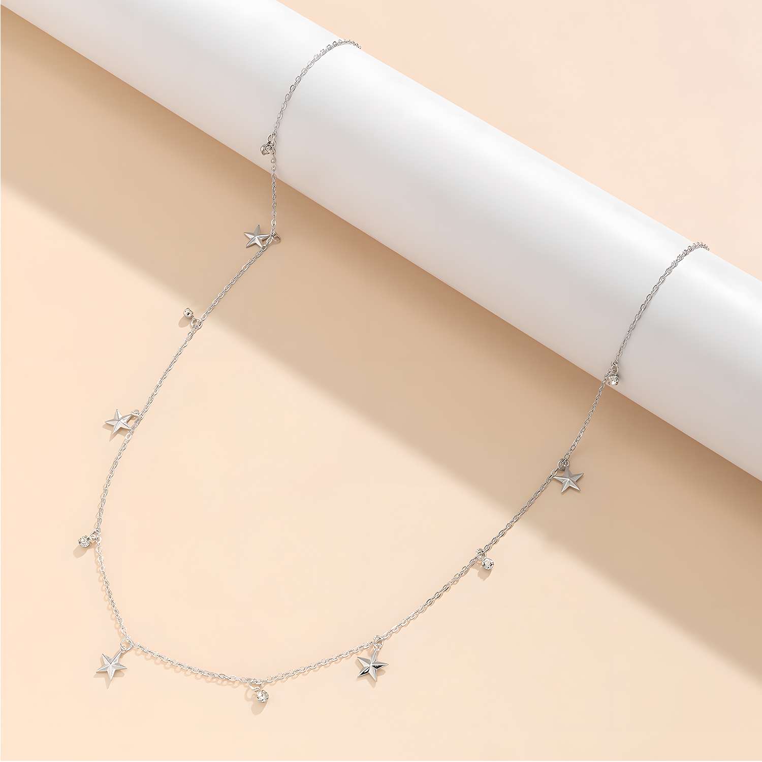 The Delicate Silver Stars Waist Chain Necklace features evenly spaced small star and heart charms on a silver chain, elegantly draped over a white cylindrical rod against a beige background.