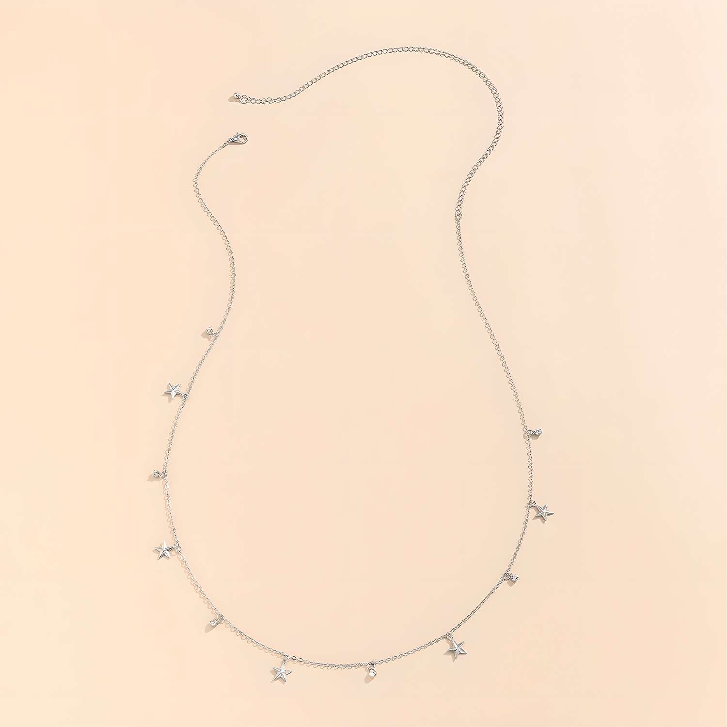 The Delicate Silver Stars Waist Chain Necklace features small star and teardrop charms on an adjustable silver chain, displayed on a light beige background. Ideal for styling with crop tops or your favorite bikini.