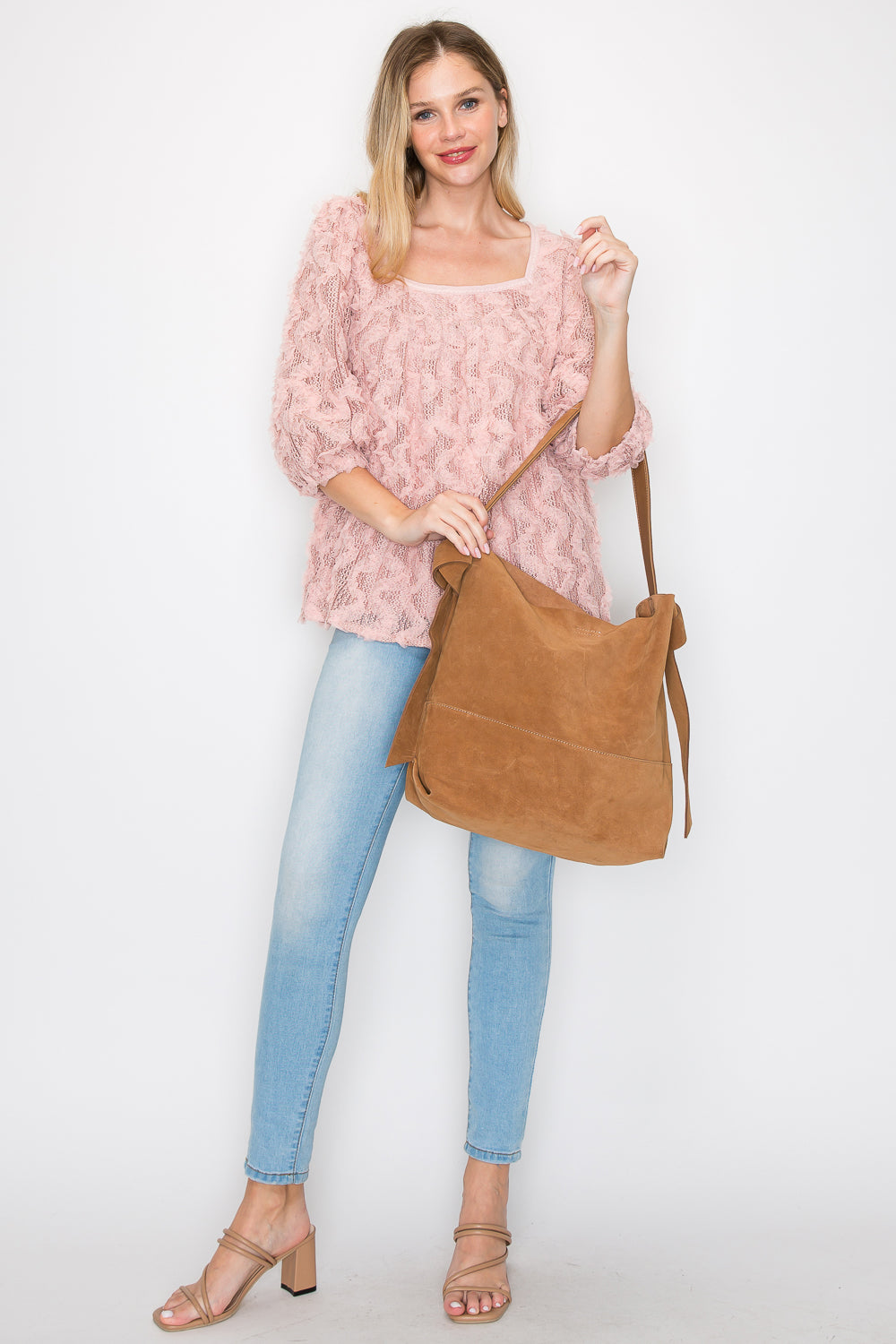 A woman smiles against a plain white background, holding the Genuine Leather Two-In-One Hobo Handbag Set with coordinating pouch, and wearing a textured pink sweater, light blue skinny jeans, and beige heeled sandals.