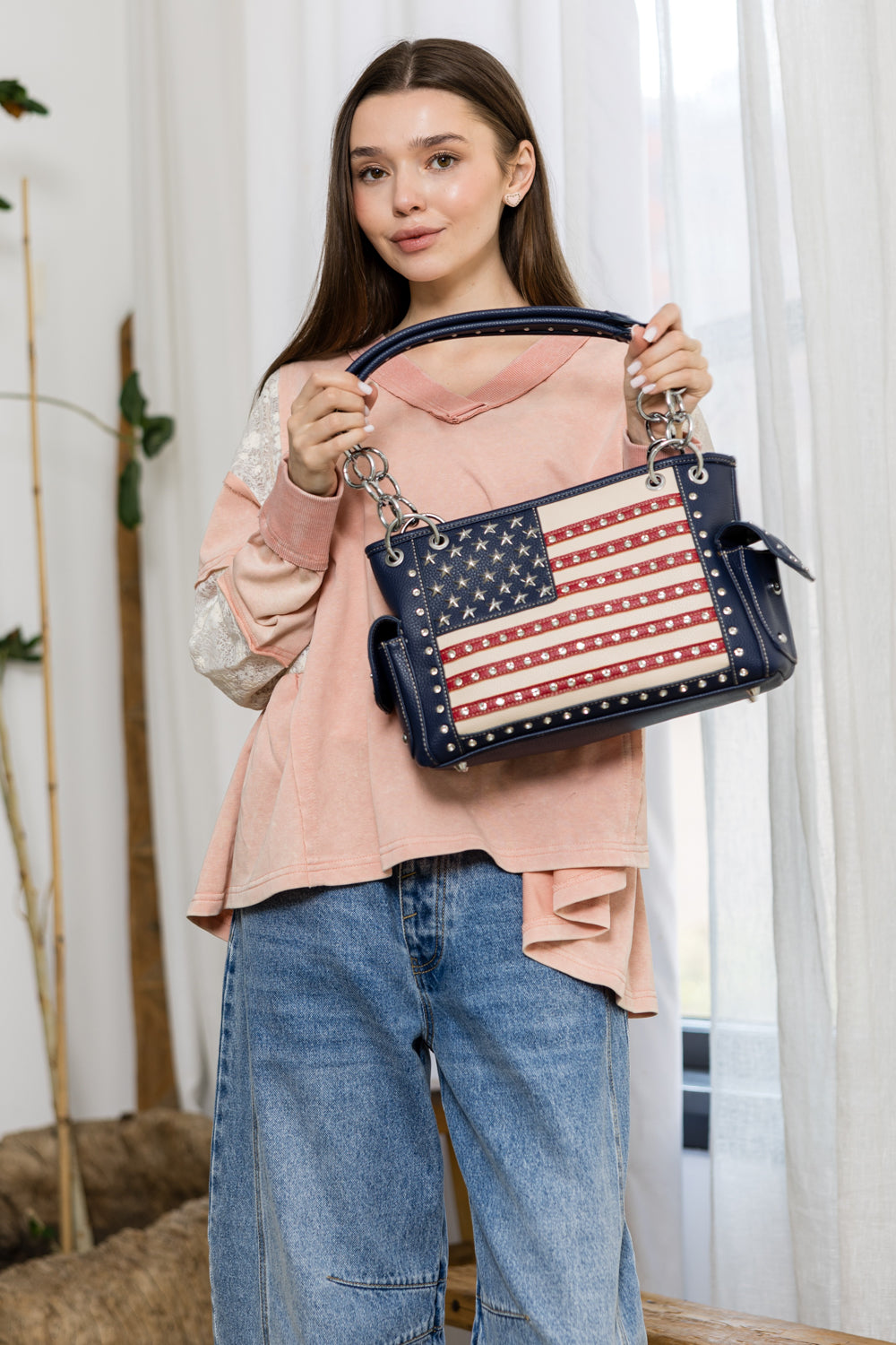 A young woman in a pink sweater and blue jeans stands indoors near a window with white curtains and a plant, holding up an American Flag Rhinestone Handbag.