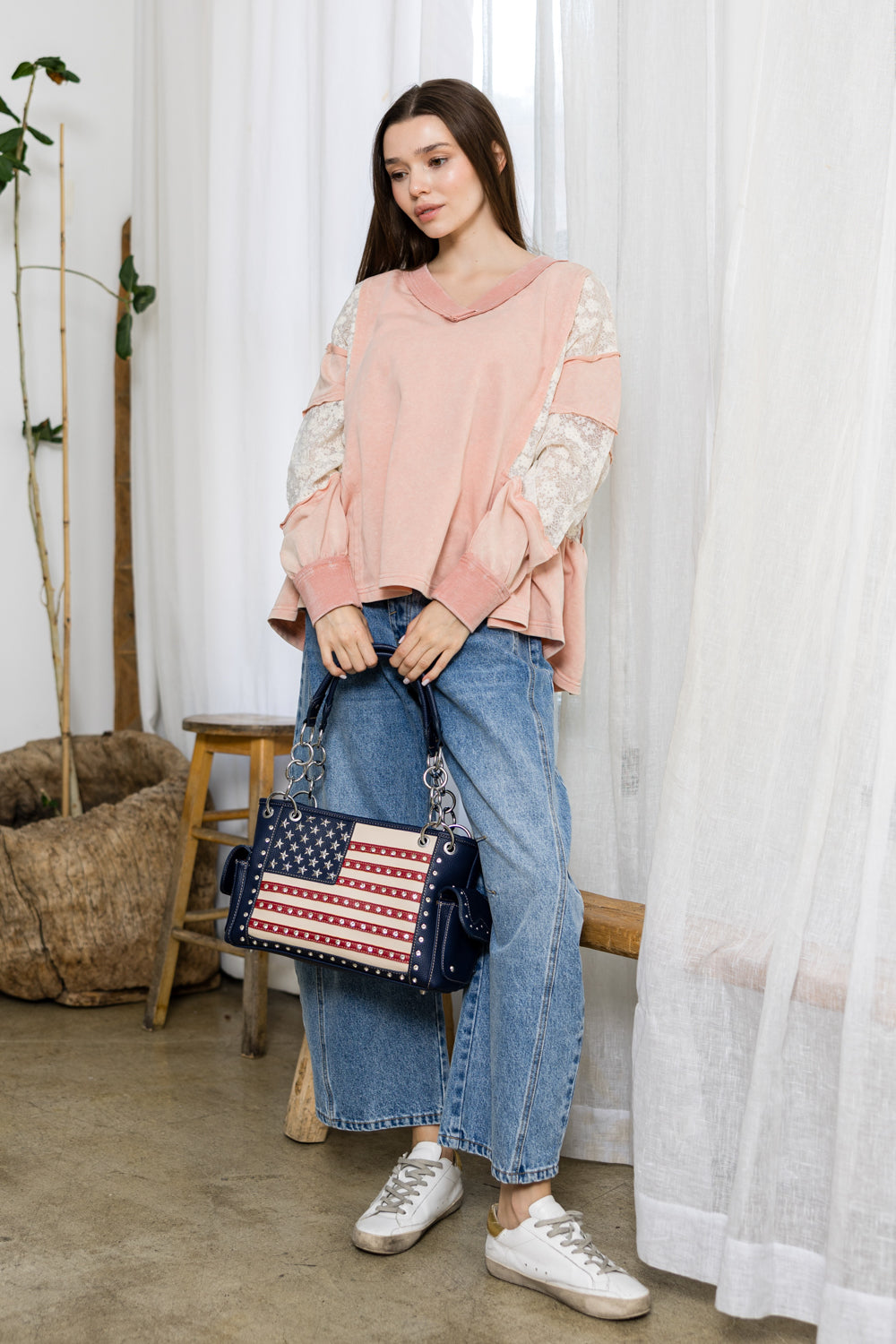A woman in a pink lace top, loose blue jeans, and white sneakers stands indoors holding the American Flag Rhinestone Handbag. She is surrounded by light curtains and rustic decor.