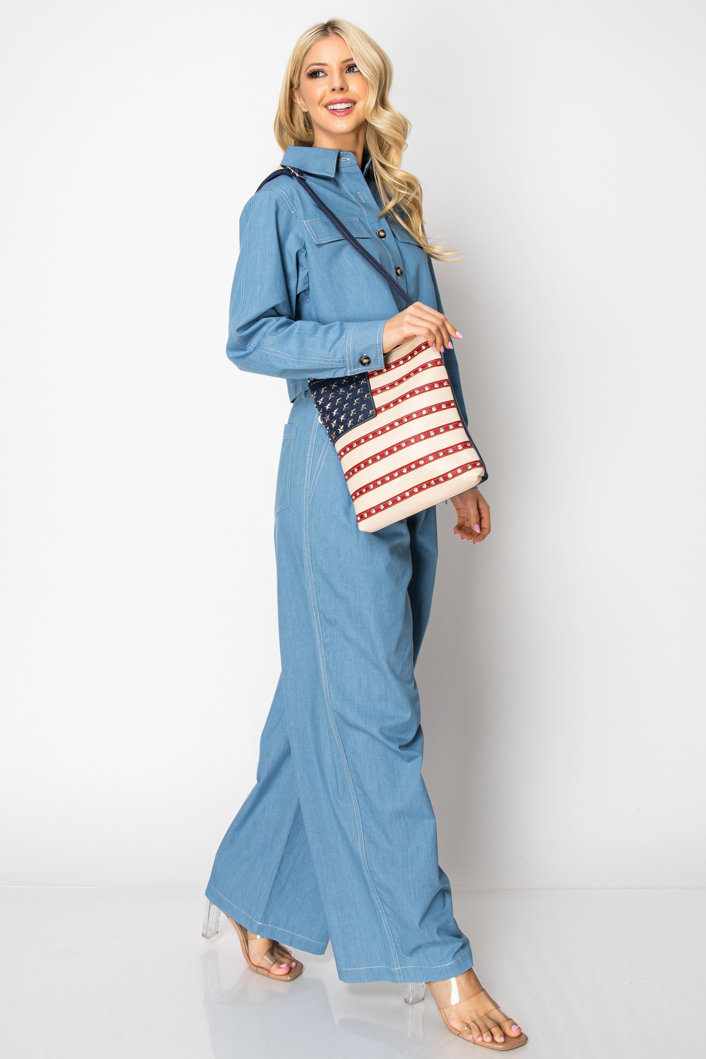 A smiling woman with long blonde hair wears a light blue denim jumpsuit and clear-heeled sandals, carrying the American Flag Rhinestone Crossbody Sling, walking against a plain white background.