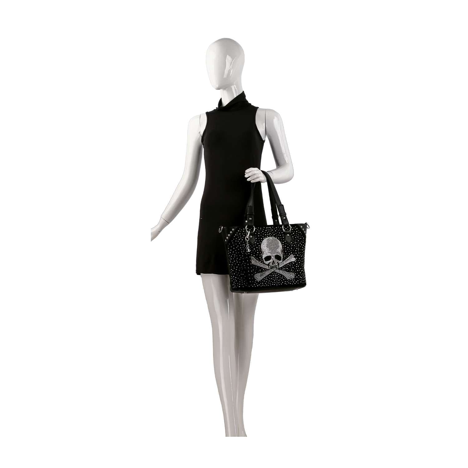 A mannequin in a sleeveless black dress holds the Rhinestone Skull Shopper Tote, featuring a bold white skull and crossbones design with polka dots—a chic faux leather shopper for standout edgy style.