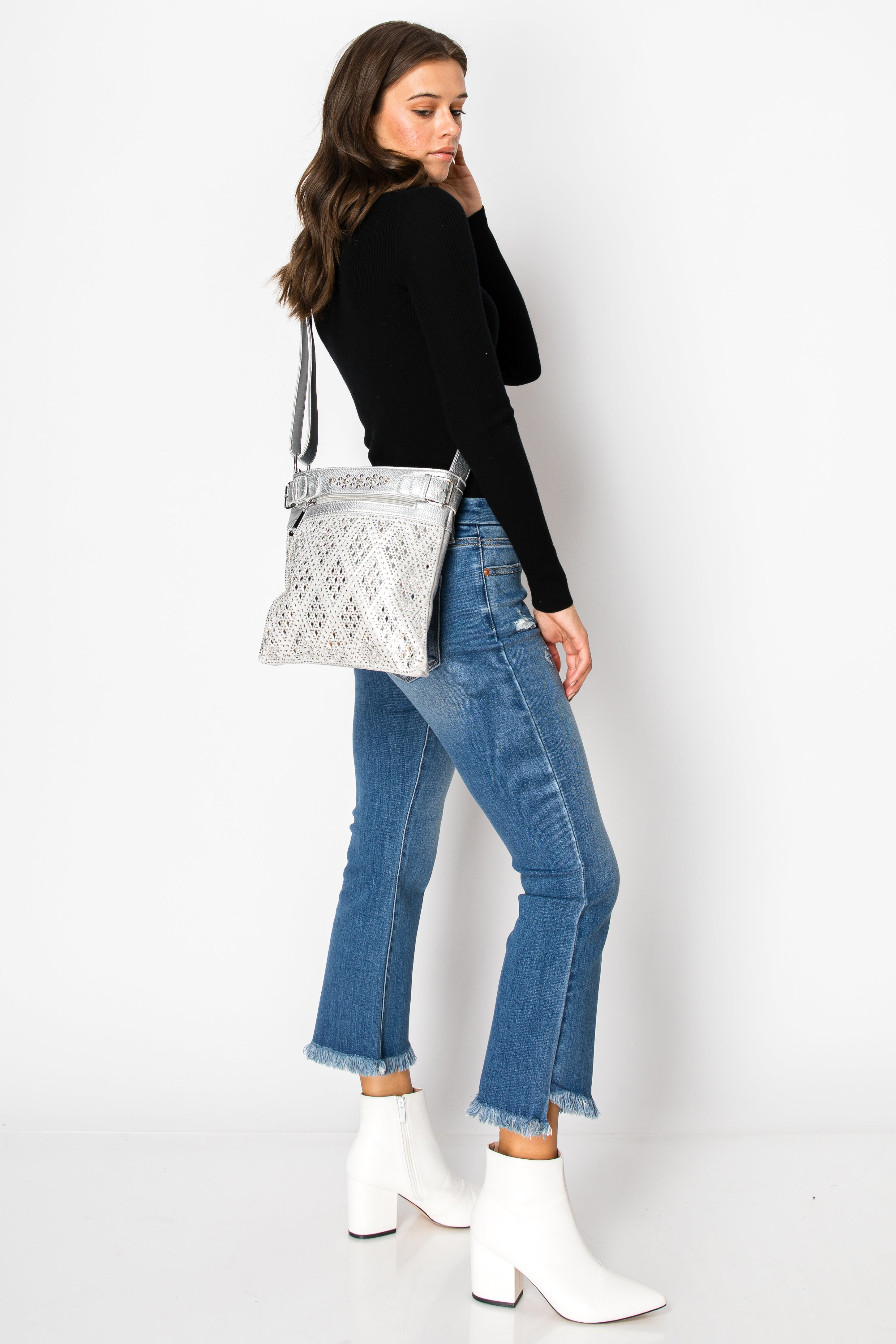 A woman poses sideways against a plain white background, wearing a black long-sleeve top, blue frayed-hem jeans, white ankle boots, and the Diamond Bling Crossbody Sling.