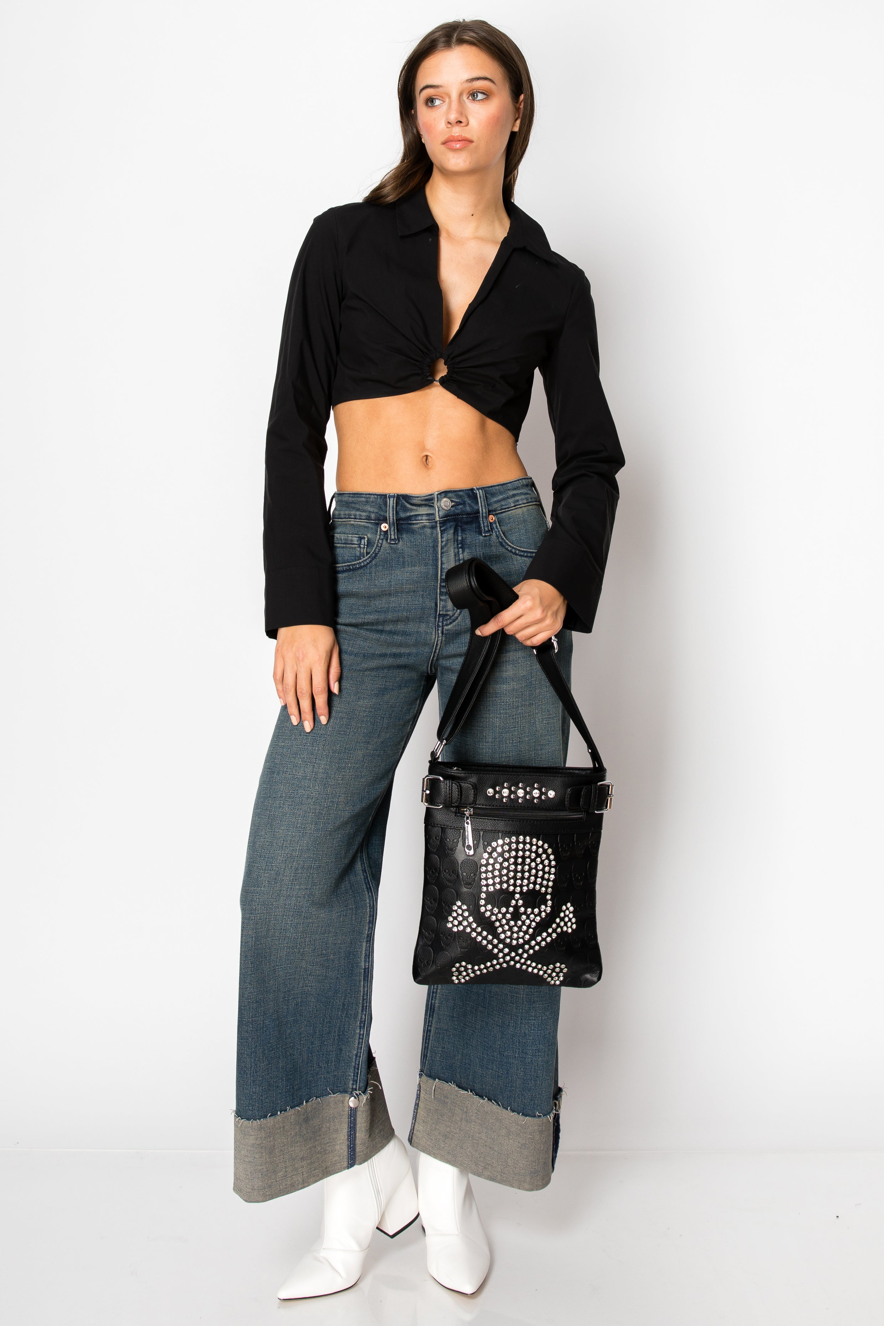 A woman stands against a white backdrop in a black cropped top, wide-leg blue jeans with gray cuffs, white boots, and carries the Spectacular Skull Design Crossbody Sling for a bold statement.