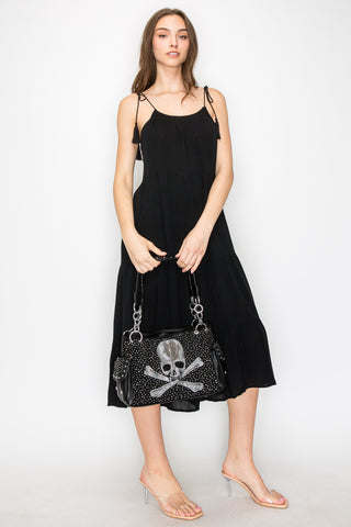 A woman in a sleeveless black dress and clear heeled sandals stands against a white background, holding the Skull and Crossbones Fashion Handbag with its striking silver rhinestone skull design.
