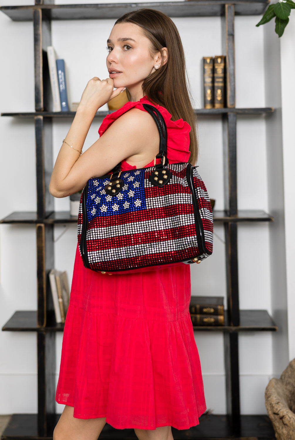 A woman in a bright red dress stands before a bookshelf, holding the Rhinestone Flag Design Locked Satchel—a faux leather purse adorned with blue, red, and silver rhinestones in an American flag pattern.
