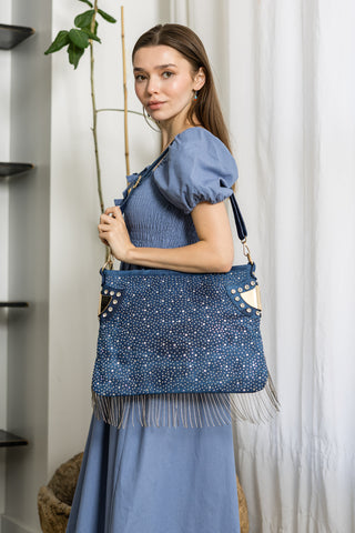 A woman in a light blue dress stands indoors, holding the Bling And Beaded Fringe Hobo Handbag over her shoulder. She faces the camera, with white curtains and a tall green plant in the background.