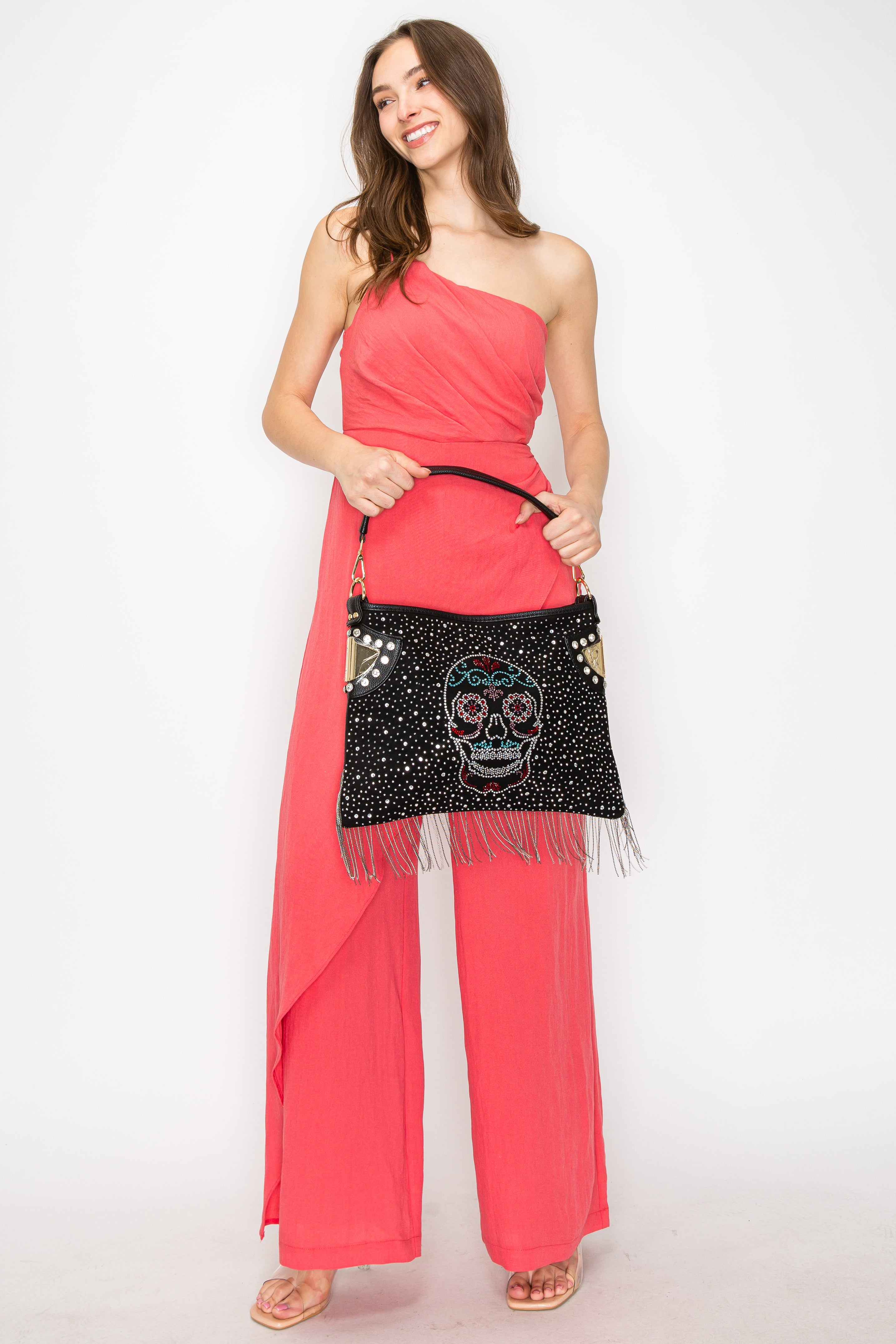 A woman in a one-shoulder coral jumpsuit and sandals smiles, holding the Sugar Skull Beaded Fringe Hobo Handbag—perfectly tablet compatible—as she poses against a plain white background.