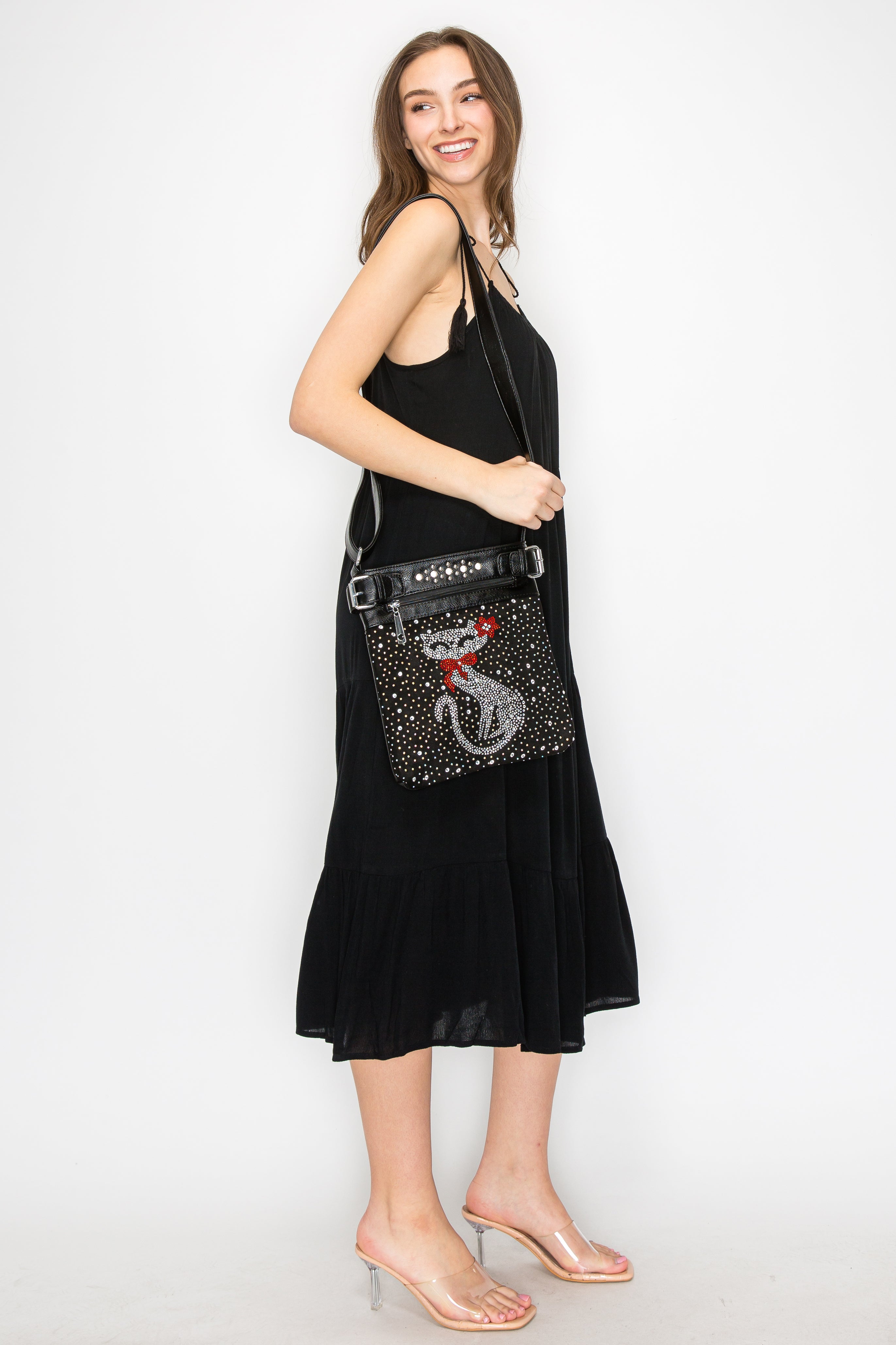 A woman in a sleeveless black dress and clear heels smiles while carrying the Cat Design Rhinestone Crossbody Sling, featuring a chic cat shape with a red bow, against a plain white background.