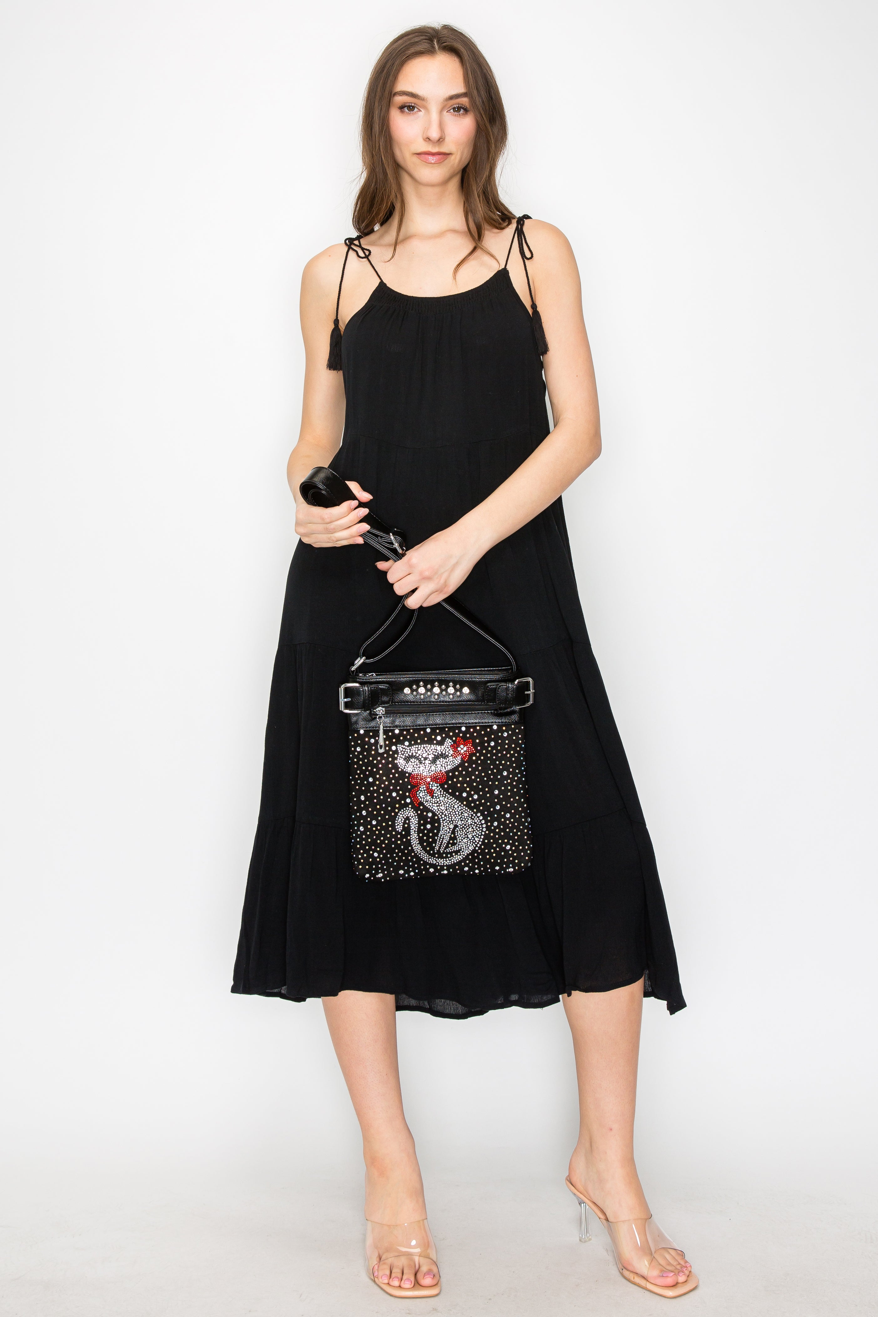 A woman stands against a white background in a sleeveless black midi dress and clear heels, holding the Cat Design Rhinestone Crossbody Sling with its sparkly cat motif.