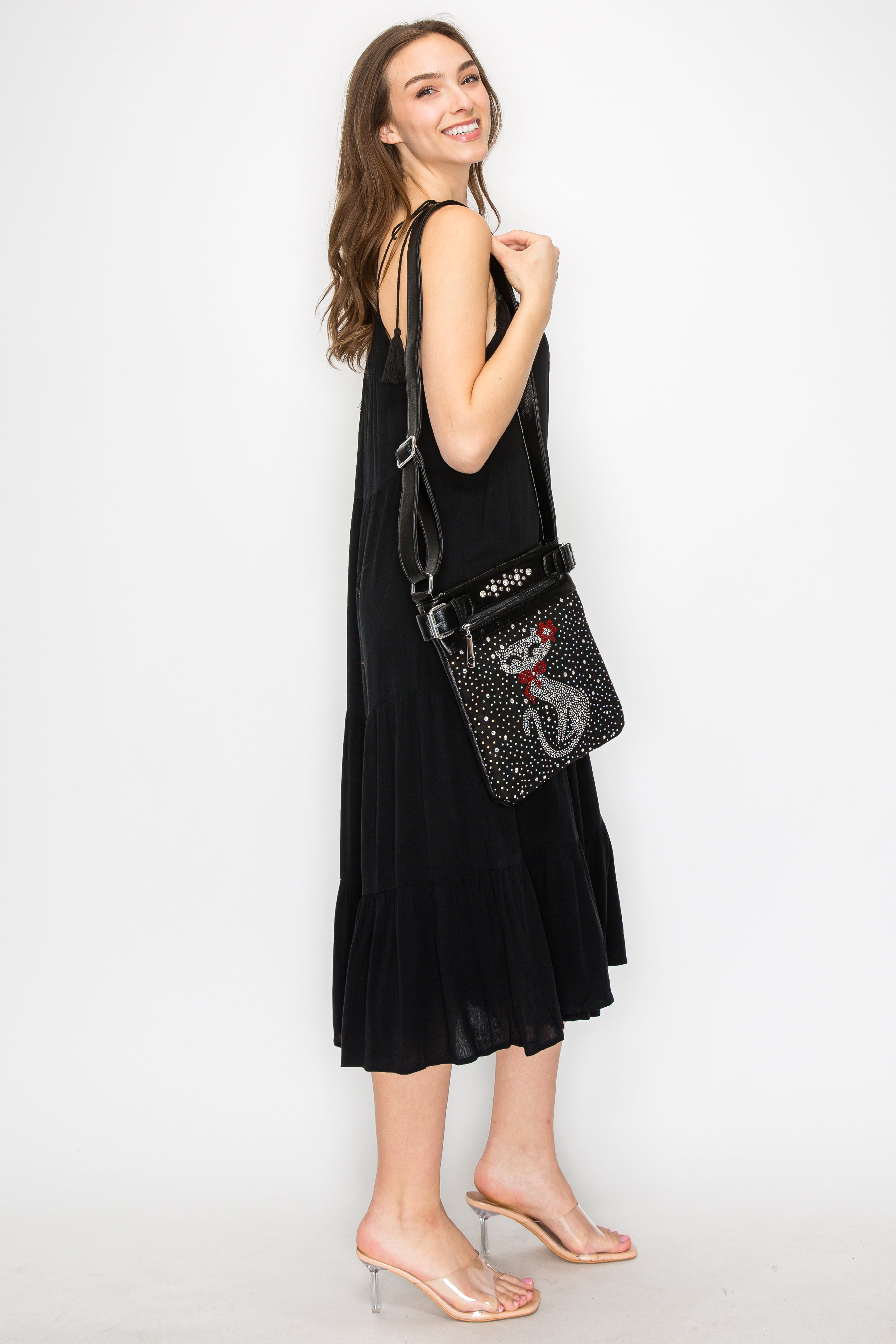 A woman with long brown hair, dressed in a sleeveless black dress and clear heeled sandals, stands smiling against a white background while carrying the Cat Design Rhinestone Crossbody Sling.