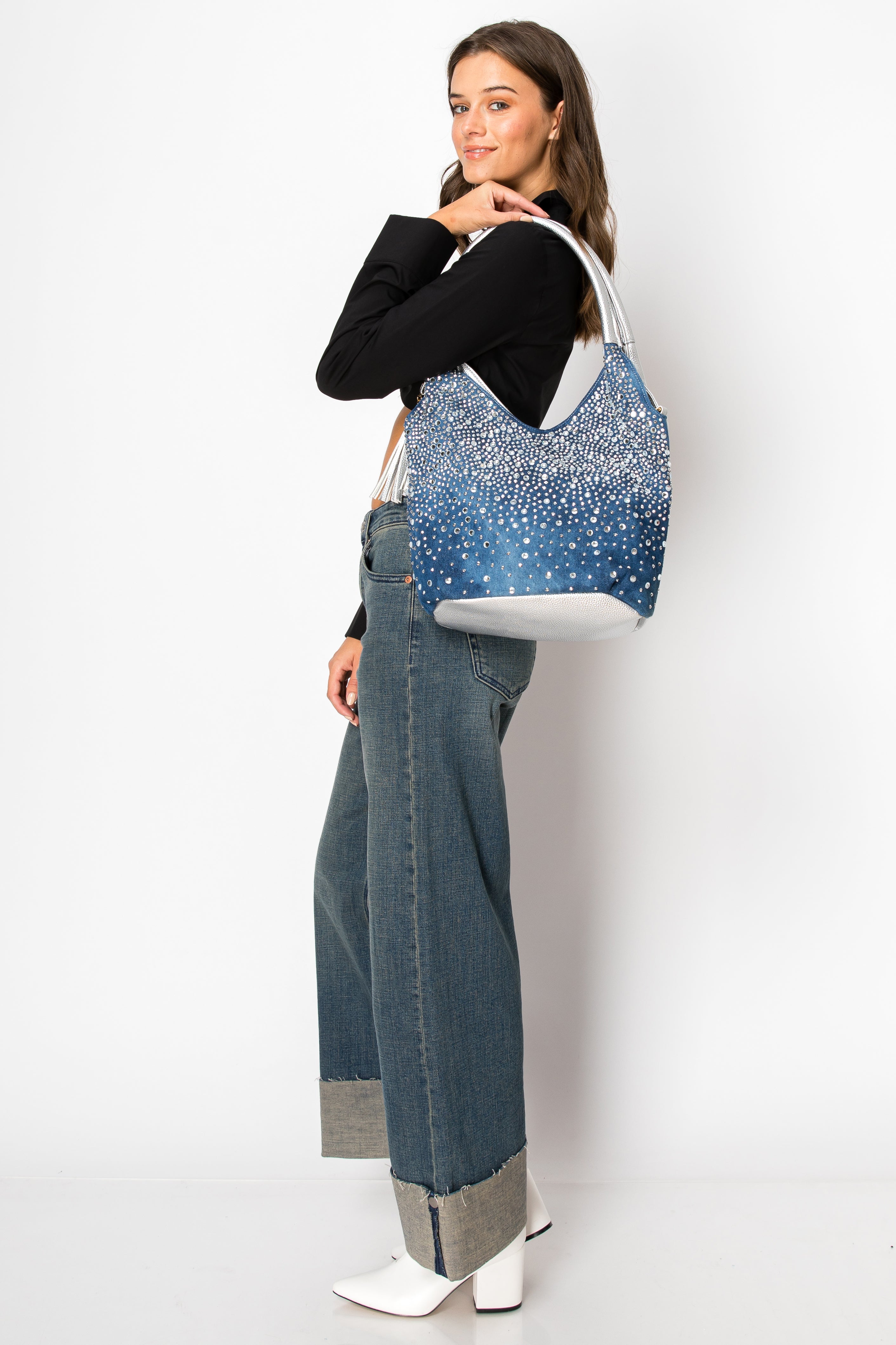 A woman wearing a black top, wide-leg cuffed jeans, and white ankle boots poses sideways with the Glamorous Rhinestone Tall Shoulder Bag against a plain white background.