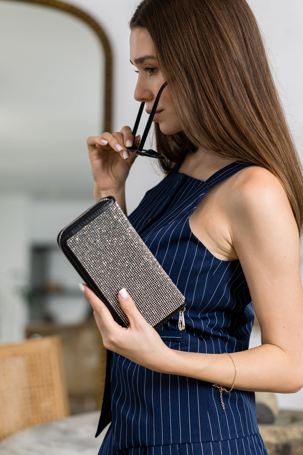 A woman in a navy pinstripe sleeveless dress holds sunglasses and the Dazzling Accordion Wallet, which sparkles with rhinestones, while standing indoors against a neutral background.