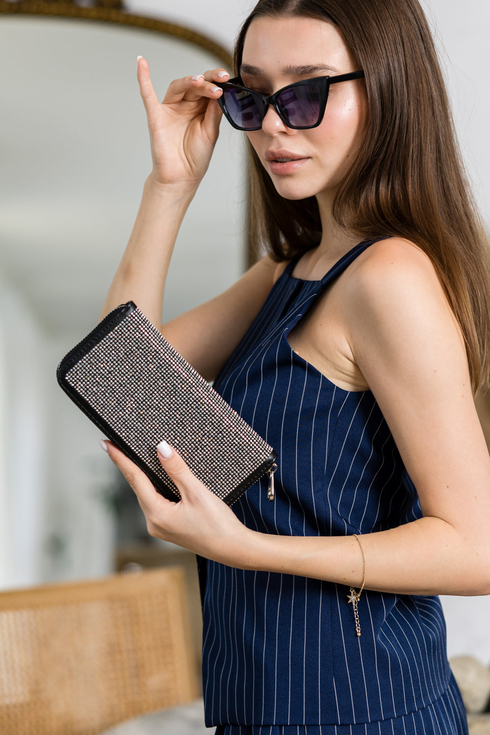 A woman wearing a navy blue pinstriped sleeveless top holds the Dazzling Accordion Wallet and adjusts her sunglasses, glancing to the side. She stands indoors, with a mirror and rattan chair in the background.