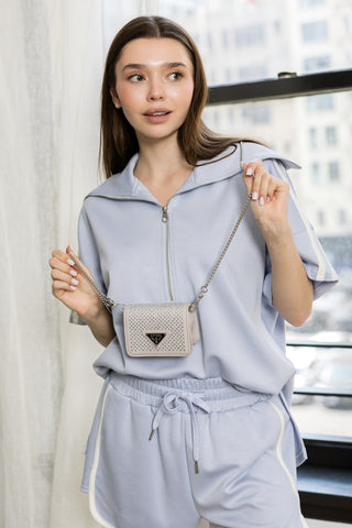 A woman in a light blue zip-up top and shorts stands indoors, holding the Petite Dazzling Front Flap Shoulder Bag Clutch with a detachable strap. City buildings are visible through the window behind her.