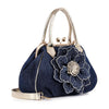 The Raised Flower Metal Frame Hand Tote features a dark blue denim body, metallic gold handles and strap, plus a bold blue floral embellishment with silver studs—making it the ideal vintage-chic statement accessory.