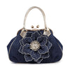 The Raised Flower Metal Frame Hand Tote is a dark blue denim handbag with metallic gold handles, featuring a large layered fabric flower adorned with rhinestones and a silver brooch at its center.