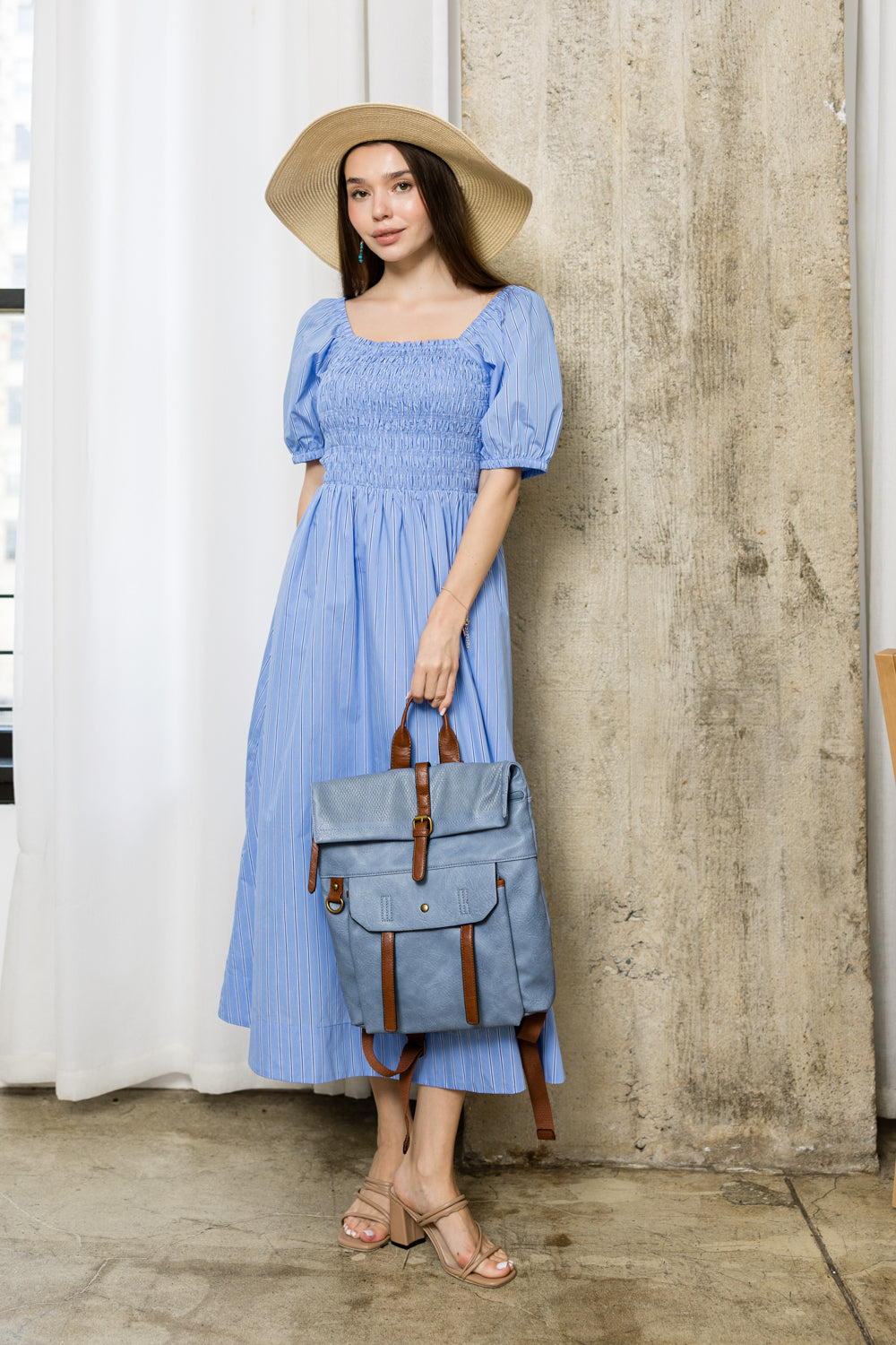 A woman in a light blue, puff-sleeve dress and wide-brimmed hat stands indoors holding the Front Pocket Fashion Backpack with adjustable straps, paired with heeled sandals near a concrete pillar.