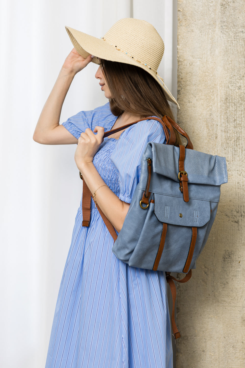 A woman in a light blue dress and straw hat stands sideways, holding her hat and carrying the Front Pocket Fashion Backpack with adjustable straps against a neutral background.