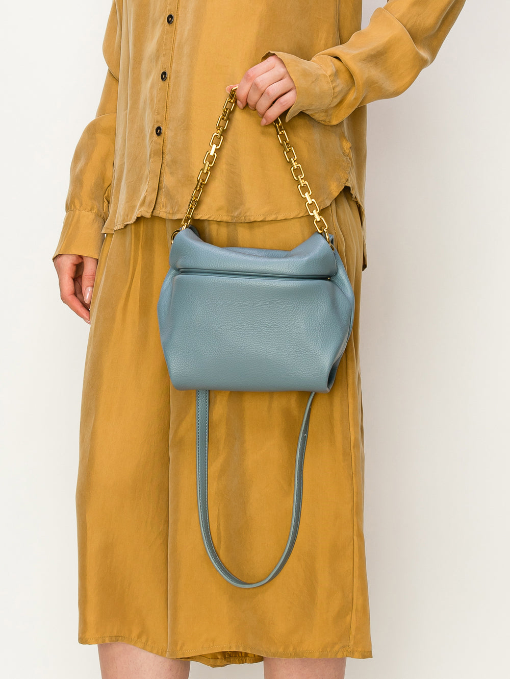 A person in a mustard yellow dress holds the Chain Accented Front Flap Shoulder Bag, a medium-sized light blue faux leather bag with gold chain and hardware, textured finish, and a long adjustable strap for versatile carrying.