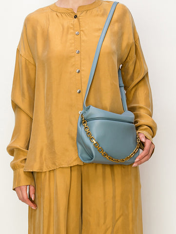 A person in a loose gold satin shirt and matching pants carries the Chain Accented Front Flap Shoulder Bag in light blue faux leather with gold hardware, worn crossbody from left shoulder to right hip.