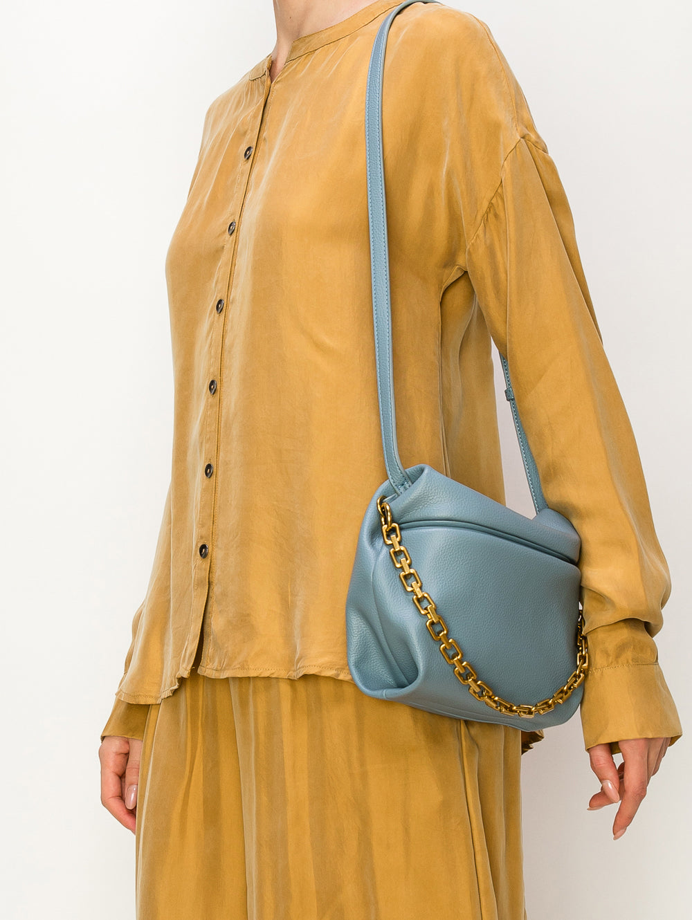 A person in a loose mustard-yellow button-up shirt and matching skirt holds the Chain Accented Front Flap Shoulder Bag in light blue faux leather with gold-toned hardware. The person's head is cropped out of the image.