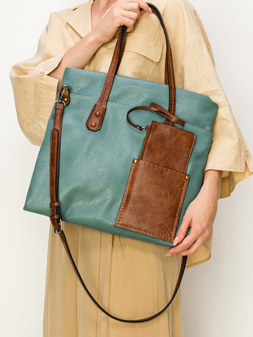 A person in beige holds the Two Toned Tall Tote Handbag, a large teal faux leather bag with brown accents and handles, tablet compatibility, a matching pocket, and a detachable shoulder strap for versatile carrying.
