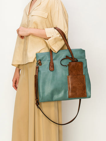 A woman in a beige shirt and tan skirt carries the Two Toned Tall Tote Handbag, a large teal faux leather bag with brown handles, front pocket, and shoulder strap. This tablet-compatible tote stands out against a plain white background.