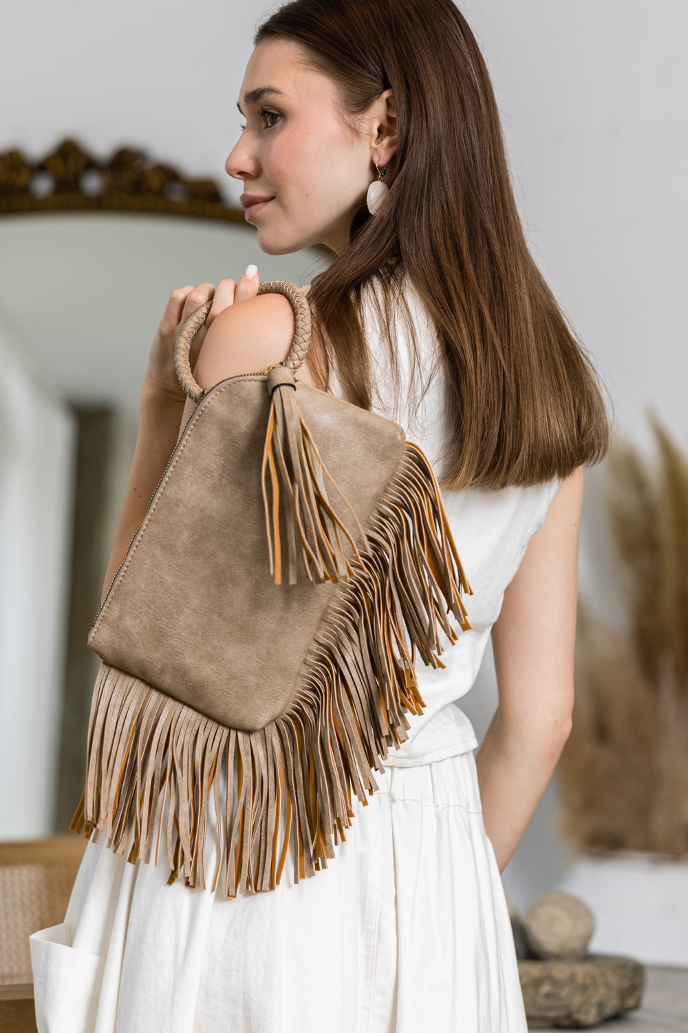 A woman with long brown hair, in a white sleeveless top and skirt, stands indoors looking to the side while carrying a beige Fringe Accented Wristlet Clutch Bag with long fringe details over her shoulder.