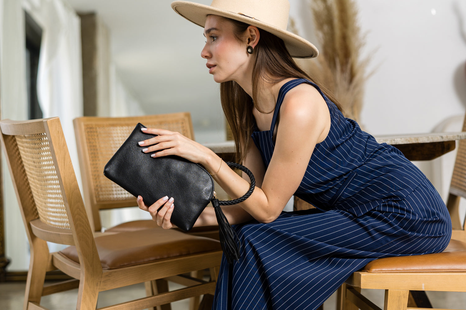 A woman in a blue pinstripe jumpsuit and wide-brimmed hat sits indoors, thoughtfully holding the Braided Handle Wristlet Clutch with credit card slots, showcasing a stylish look in a modern setting.