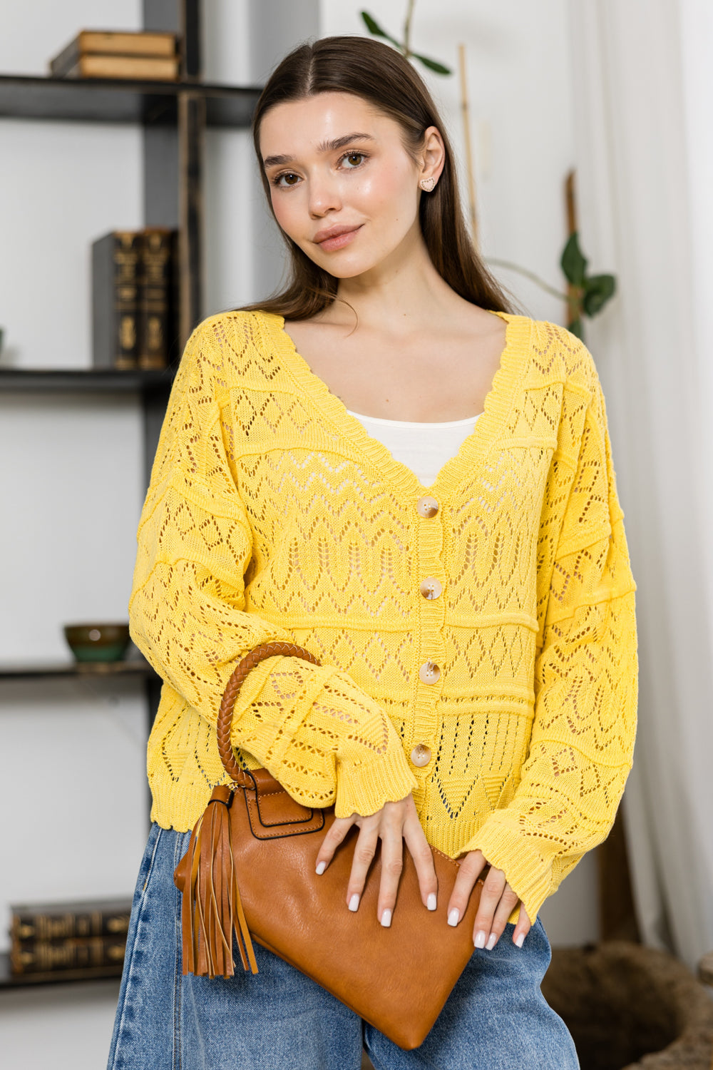 A woman in a yellow knit cardigan and blue jeans holds the Braided Handle Wristlet Clutch, a brown faux leather wallet with tassels and zip closure, smiling softly indoors in front of shelves with books and decor.