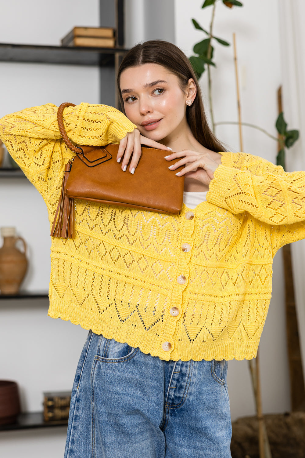 A woman in a yellow knit cardigan and blue jeans poses indoors, holding the Braided Handle Wristlet Clutch near her face with shelves and plants in the background.