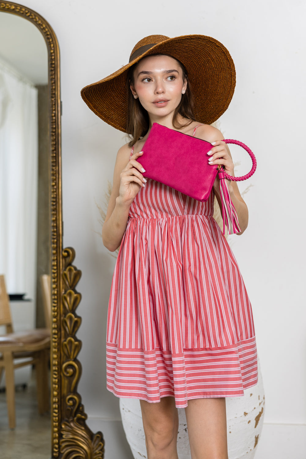 A woman in a wide-brimmed brown hat and pink striped dress holds the Braided Handle Wristlet Clutch, standing indoors near an ornate mirror and white decorative vase.