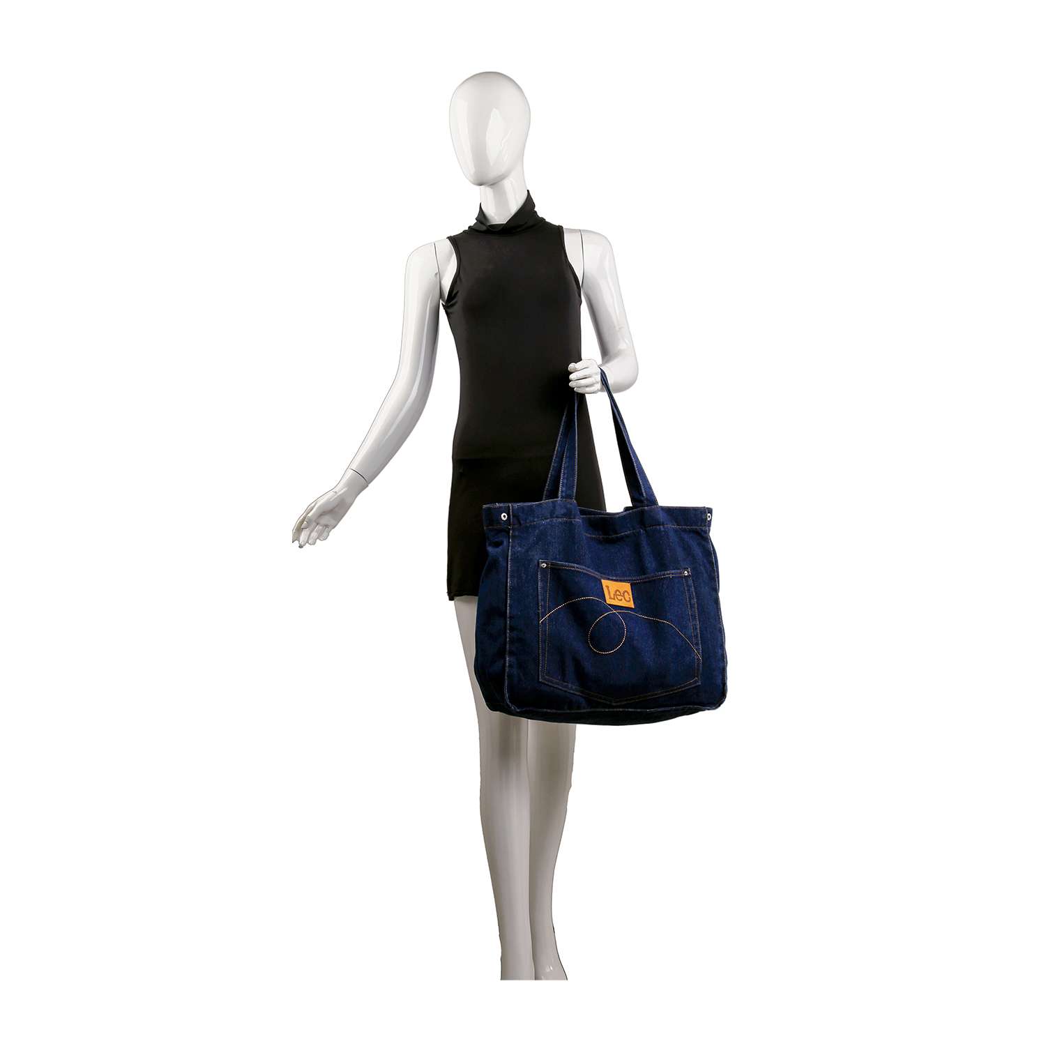 A mannequin in a sleeveless black dress holds the Front Pocket Large Denim Tote—a spacious blue denim bag with front pockets, tan patch, and double handles—standing against a white background.