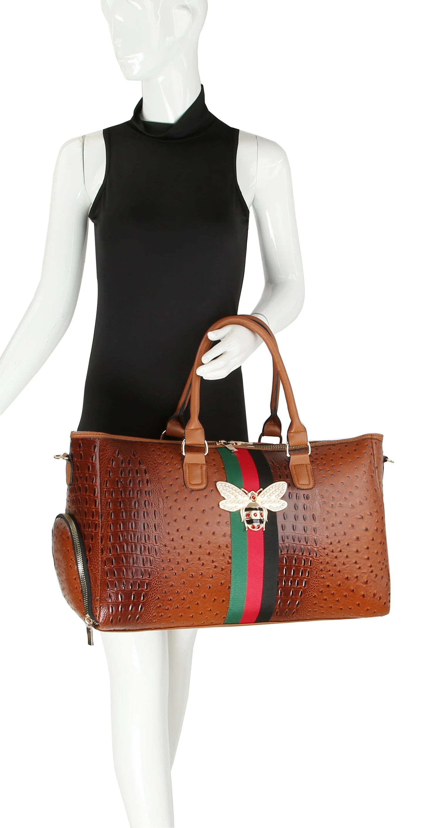 A mannequin in a sleeveless black dress holds the Croc Embossed Rhinestone Bee Weekender, a large brown faux leather handbag with a green and red stripe, rhinestone bee emblem, and gold-toned hardware.