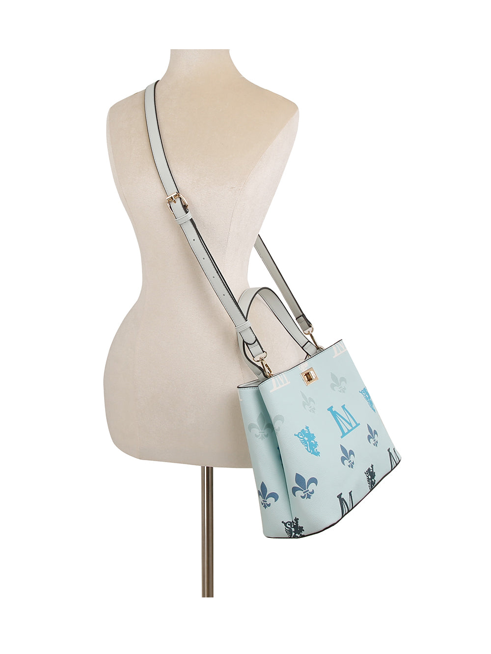The Monogram Print Lux Faux Leather handbag, shown on a dress form, features blue and navy fleur-de-lis motifs, gold-toned hardware, top handles, a wide shoulder strap, and is tablet compatible.