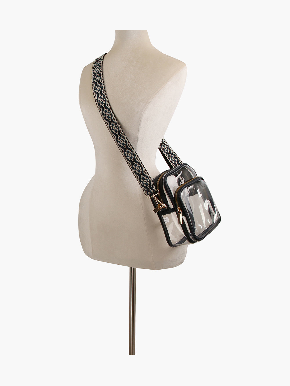 A dress form mannequin displays the Guitar Strap Clear Crossbody Bag, a boxy clear bag with black trim, gold hardware, two zippered compartments, and a wide patterned shoulder strap worn diagonally across the torso.