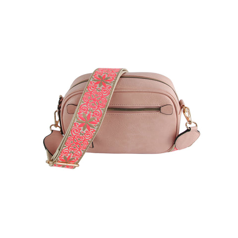 The Guitar Strap Crossbody Sling is a blush pink faux leather bag with two zippered compartments, gold-tone hardware, and a wide detachable strap adorned with a pink and gold floral pattern.