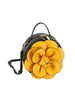 The Raised Flower Petite Handbag is a round black faux leather bag featuring a large decorative yellow flower, gold-tone hardware, zip closure, top handle, and an adjustable black shoulder strap.