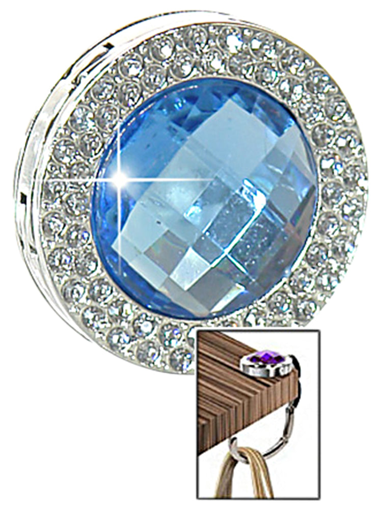 The Gem and Rhinestone Purse Hanger is a round, silver accessory with a faceted blue gem center and rhinestones around it, shown holding a handbag from the table edge.