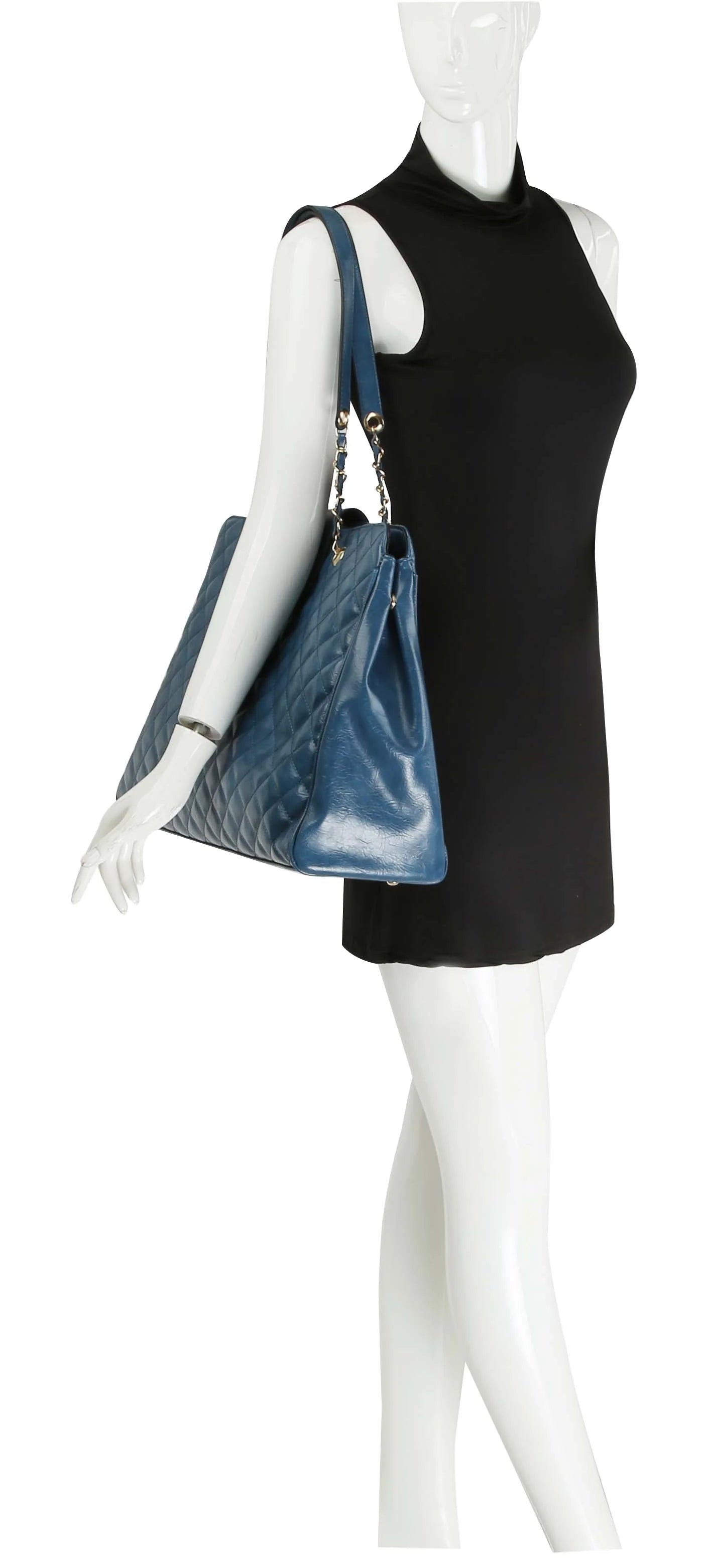 A mannequin in a sleeveless black dress showcases the Oversized Quilted Tote Travel Bag, a large blue faux leather shoulder bag with chain and leather straps.